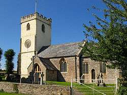 Stone building with lighter coloured square tower. In the foreground are gravestones.