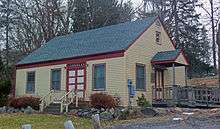 A smaller building with a pointed roof in the same colors as the meeting house. It has wooden steps in the front at the center and a wooden ramp at the right