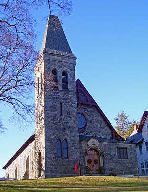 A triangular stone building with a peaked-roofed tower attached to its front slightly to the left, illuminated by the sun from that direction. Its wooden doors have a Christmas wreath on them