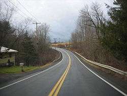 Ground-level view of a two-lane road passing through a rural area, with only one home visible on the left-hand side. Wooded terrain opens to a field as the highway curves towards the left.