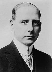 Back and white photograph of a white man wearing a high collared shirt, tie, and dark jacket