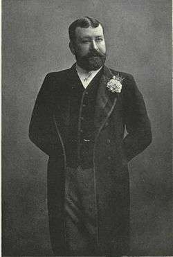 G. R. Sims, a dapper paunchy gentleman with a black beard and hair wearing smart clothes and a buttonhole