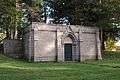 A wide, rectangular, brownstone structure with many gothic elements is seen, including small buttresses. From behind the structure is surrounded by trees. Door is aged metal.