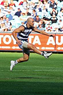 Young male athlete is in a navy blue and white striped sleeveless shirt and blue navy shorts. He has both feet off of the ground as he kicks the red football and watches the follow-through.