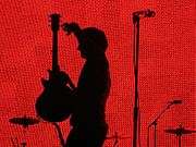 A man standing in front of a red background with a guitar in his hand.