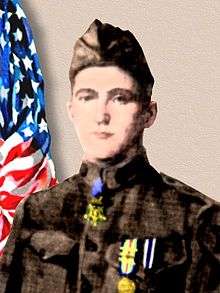 Head and shoulders of a young man in a brown military uniform with a garrison cap. A medal hangs down from his neck, the strap lying under his collar, and two medals hang from colorful ribbons on his chest.