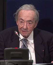 A head-and-shoulders photograph of an elderly man standing at a lectern and speaking