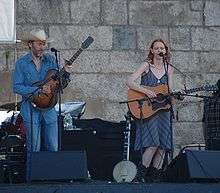 A slender, bearded, middle-aged man in a blue shirt and jeans and Welch in a black dress playing guitar on stage. Welch is singing.