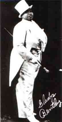 A publicity photo of a stout African American woman in white tuxedo with tails and top hat, carrying a cane and her signature in the lower right corner