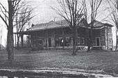 A house of wooden construction with a wrap-around front porch and trees in the front yard