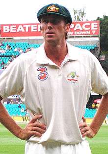 A man in cricket whites and a baggy green cap, with his hands on his hips. He is standing on the pitch in front of a stand with a number of empty turquoise seats.