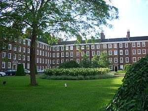  A grassy foreground, with a tall tree and shrubs, with a terrace of red brick buildings in the background and left side