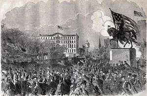 Crowd surrounding an equestrian statue topped by a huge U.S. flag.