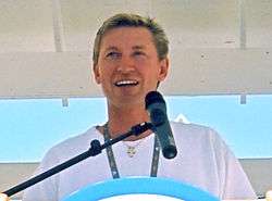 Upper body of a man with dirty-blonde hair who is looking into the distance. He is standing at a podium and speaking into a microphone.