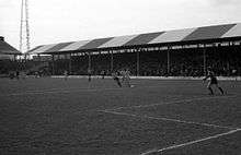 A black-and-white photograph of a football match in progress in front of a modest, single-tiered, British-style grandstand