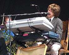 Colour photograph of Guillemots lead singer Fyfe Dangerfield performing live in 2006. He is sat behind a keyboard and singing into a microphone.