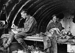 Two men in military uniform in a building. The man on the left has something in his mouth and is sitting down looking at some papers, angled away from the camera. The men on the right is smiling and looking at the camera. The roof of the structure is curved.