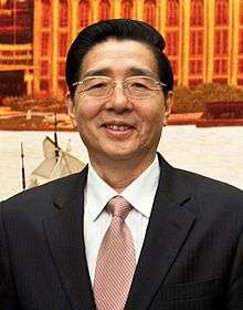 a smiling, clearly aged man with wavy haircut, wearing glasses, a white shirt, a suit and a light red tie