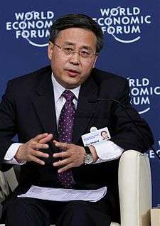 a serious looking man bending forwards while seated, wearing glasses, a white shirt, a suit and a purple tie with white dots