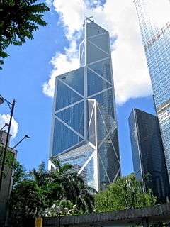 A tall tower coated with reflective glass and steel X patterns rises over trees and smaller buildings.