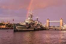 The bow of a large blue warship, moored on a river, with a bridge in the background.