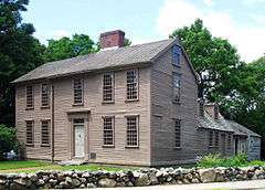 The main part of the home is a wooden,  2 1⁄2-story rectangular building with large windows, one central door, and a central chimney. A smaller wing extends back from the right side. There are large trees in the background and a low rock wall in the foreground.