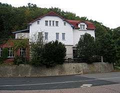 A road and behind it, at a somewhat higher level, a large, three-storey white house with a red roof; a wooded hill is in the background. The main house has an extension to the left, and a separate three-storey wing leading off to the right; the colour of the road surface changes abruptly at the point where the main house ends and the wing to the right begins.