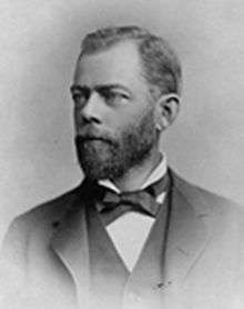 Head and shoulders of a white man with a full beard, wearing a dark suit coat, vest, and bow tie.