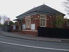 A small single-storey red brick building with cream stone window surrounds, mullions and transoms. An awning projects above the doorway and a slated pitched roof is topped with two finials.