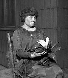 A woman with full dark hair and wearing a long dark dress, her face in partial profile, sits in a simple wooden chair. A locket hangs from a slender chain around her neck; in her hands is a magnolia, its large white flower surrounded by dark leaves.