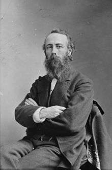 A white man with a scraggly beard and wearing a suit, sitting in a chair with his arms crossed.