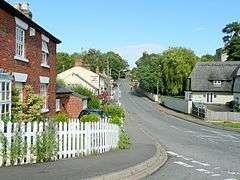 A red brick house in the foreground. Further down the street is a thatched house, behind which the church tower can just be seen, and across the road is a pub.