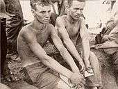 Two men without shirts on sit surrounded by soldiers