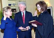 Hillary Clinton taking oath as Secretary of State on January 21, 2009. She is on the left side of the image, facing toward the right. The oath is being administered by Associate Judge Kathryn Oberly, who is standing directly in front of Hillary (on the right side of the photo) and facing toward the left. Bill Clinton, who is standing on both women's side in the background of the image, is holding a Bible.