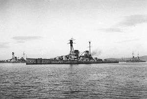 Side view of a large warship with a prominent tripod mast above the superstructure at anchor. Four gun turrets and two funnels are visible.