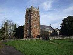 Stone building with prominent three stage square tower. In the foreground is a grass area and road separated from the church by a stone wall.