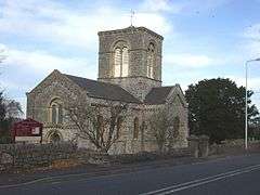 Stone building with arched windows and central square tower.In the foreground are trees and a road.