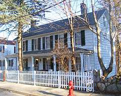 A white wooden house with full front porch, black window shutters and chimneys at either end behind a white picket fence and some bare trees, lit by sunlight from the right