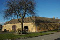 The Jubilee Tree stands on a small patch of grass, the tree is surrounded by a bench and behind it is an old farm building terraced with other houses.