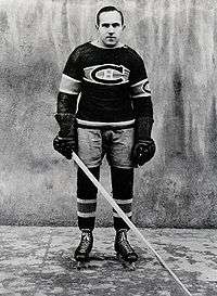 The ice hockey player stands, wearing a Canadians jersey with the traditional large "C" crest and big leather gloves and other older style equipment.