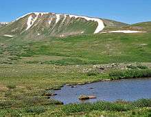 A grassy yet barren landscape with a small pond in the right foreground and distant ridgeline and mountains in the background