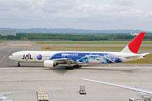 A Boeing 777–300 aircraft with special Oneworld livery taxiing from the tarmac on to the taxiway, with a mountain view on the background