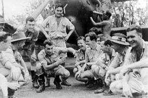 Eleven young men crouching or standing in front of a propeller-driven aircraft which another man is working on. The crouching man in the centre of the photograph is gesturing with his hands and the men around him are laughing.