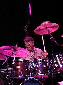 A man in a striped dress shirt, sitting behind and performing on a drum set