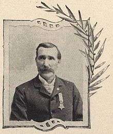 Head and shoulders of a white man with a mustache, wearing a suit coat with a star-shaped medal pinned to the left breast, over a white shirt.