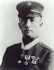 Head and shoulders of a white man in military uniform with a peaked cap, a medal around his neck and one medal, two ribbon bars, and a badge on his left breast.