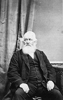 John Cracroft Wilson sitting on a chair, visible from above the lower leg, wearing medals pinned to his jacket, with a full white beard.