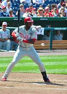 A dark-skinned man wearing a gray baseball jersey and pants with red trim and a red batting helmet and holding a baseball bat