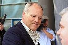 A grey-haired, bald man wearing a blue jacket and wearing an open-collar white shirt with blue stipes. His body is side-on and is head is turned in the direction of the camera.