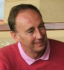 A smiling middle-aged white man with short hair, wearing a pink shirt and red pullover, looking to his left with the tip of his tongue between his lips